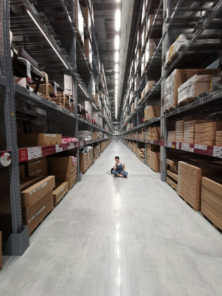 An Asian woman sits in a large warehouse aisle filled with inventory racks and shelves.